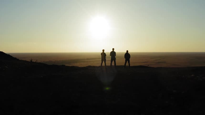 The silhouettes of a trio of men standing atop a mountain while looking out at the vast countryside below