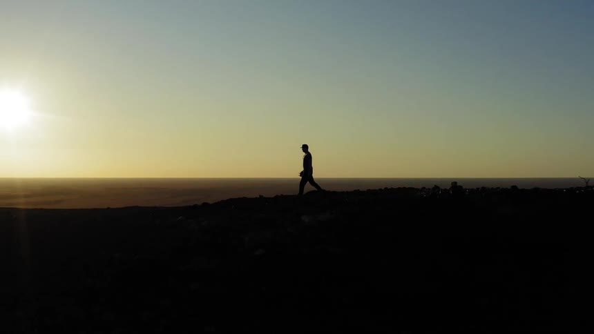 The silhouette of a man walking along a cliff overlooking a countryside while carrying a pair of binoculars at sunset