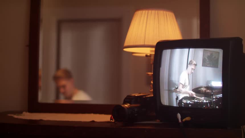 A man plays the drums while recording himself with a video camera and displaying the recording on a television screen