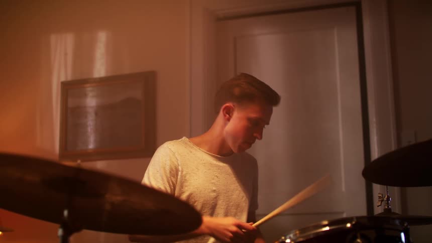 A young man rhythmically strikes a drum set with a drum stick while sitting in a dimly lit room