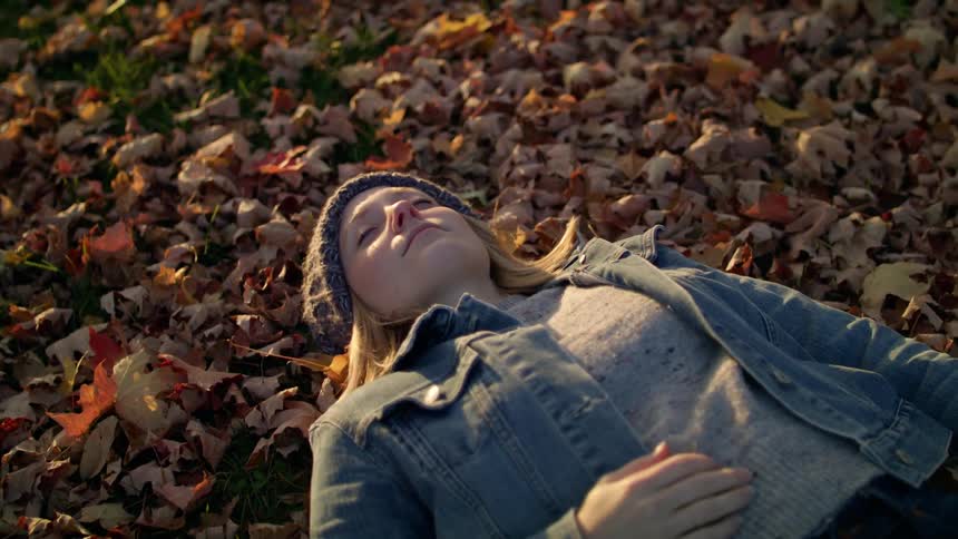 Blonde woman with a beanie and a denim jacket lies on brown leaves in the forest