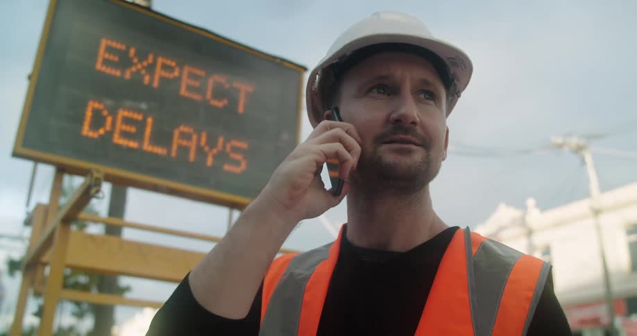 A construction worker talks on a cell phone standing outside in front of a large electronic road sign