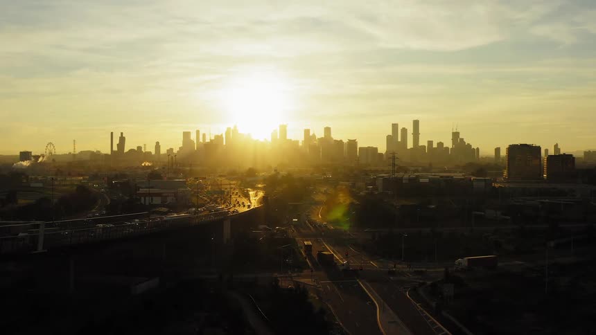 Sun shines in cloudy morning sky behind city skyline as traffic flows along busy highway