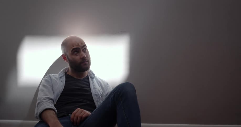 Bald man with a beard sits down and leans against the wall in a room