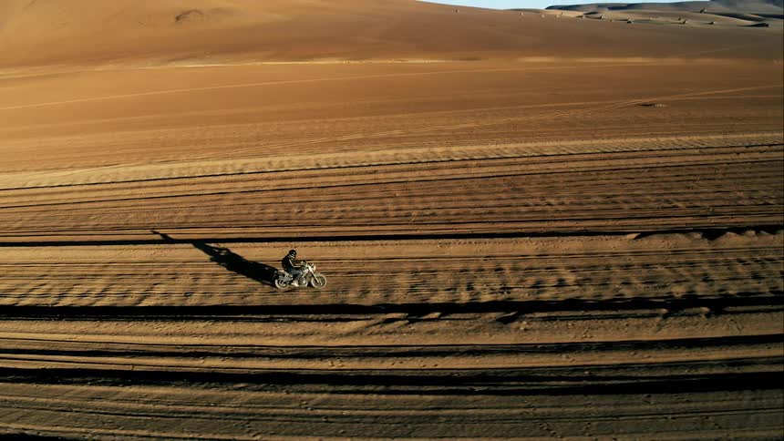 Aerial view of man riding a motorcycle through the desert near the mountains at sunset