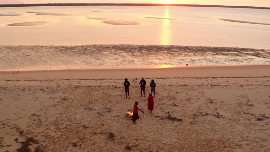 Three men clap as two women dance around a fire on a beach at sunset