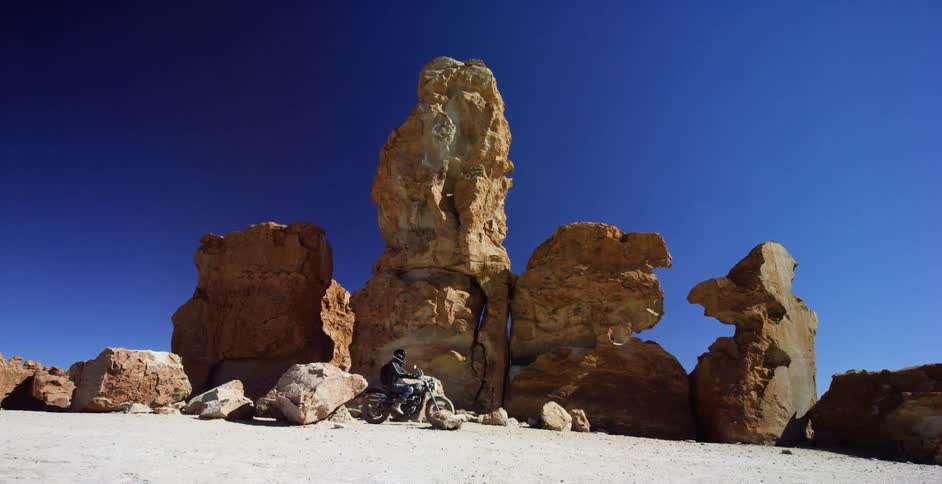A biker rides a motorcycle off road base a large rock formation in the desert under a clear daytime sky