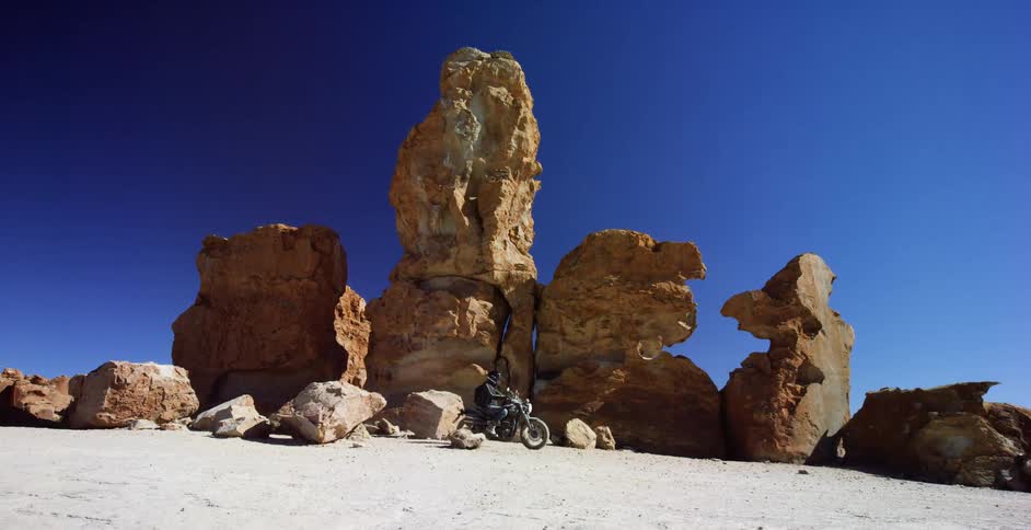 A biker rides a motorcycle offroad past a rock formation in the desert under a clear blue sky