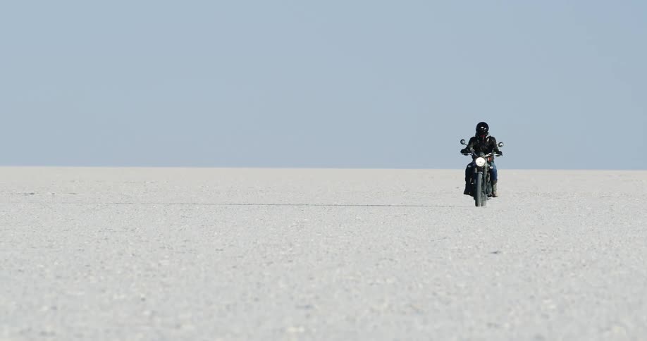 Motorcyclist with a helmet a black leather jacket and jeans rides a motorcycle across the large white sand desert