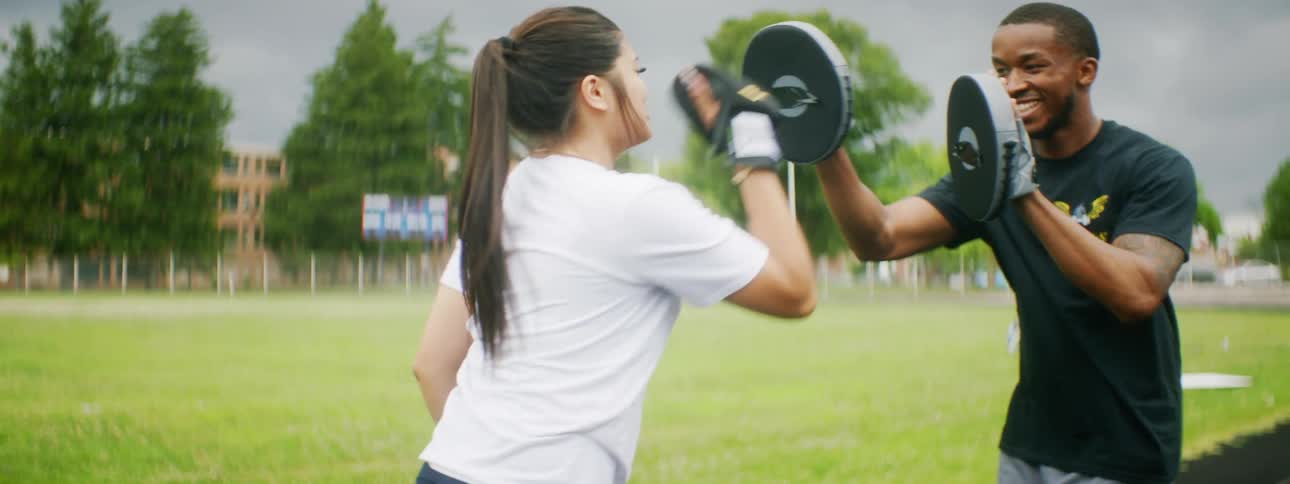 Man holding strike pads for woman in boxing gloves on field outdoors