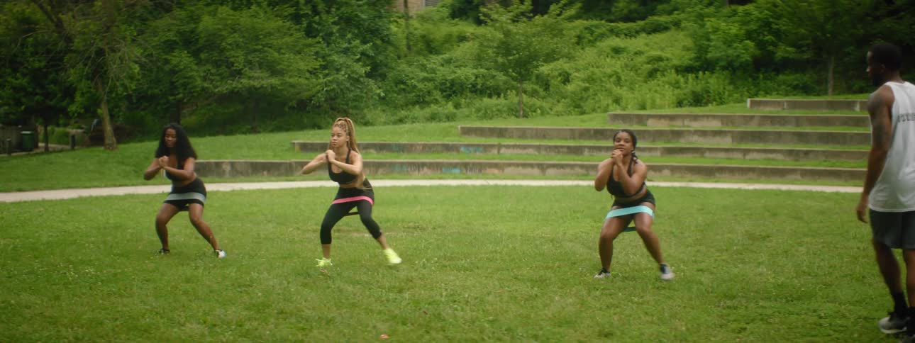 A man instructs three young women as they exercise with resistance bands around their thighs in a park during the day