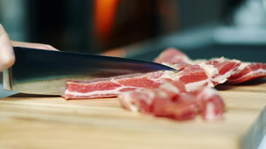 A cook uses a kitchen knife to slice bacon on a wooden cutting board