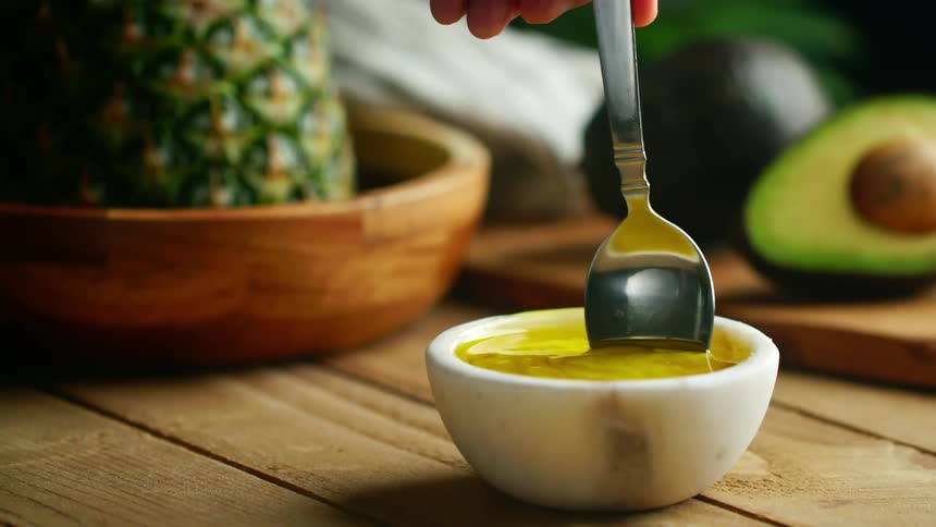 A person with a spoon mixing a yellow liquid in a bowl with avocados and a pineapple in the background