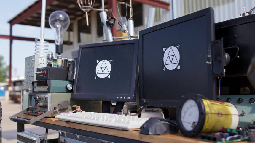 Two monitors of a computer showing a strange set of symbols on a desk with other electronic equipment and light bulbs