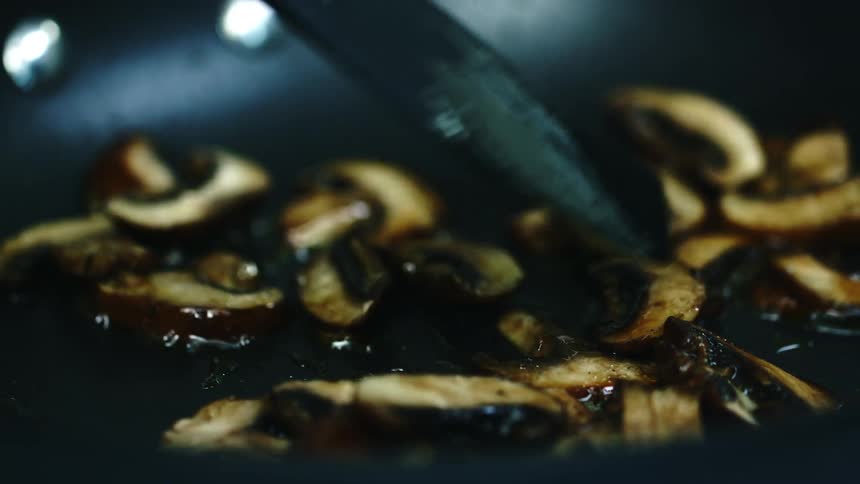 Black spatula with a wooden handle moves mushrooms in a sauce around a black pan