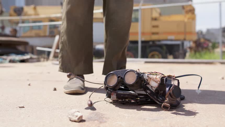 A person with gloves with wires walking up to goggles and picking it up with a fence in the background