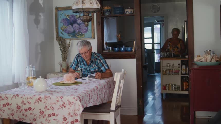 A man sitting at a table reading a book while a woman in the background is cooking in the kitchen