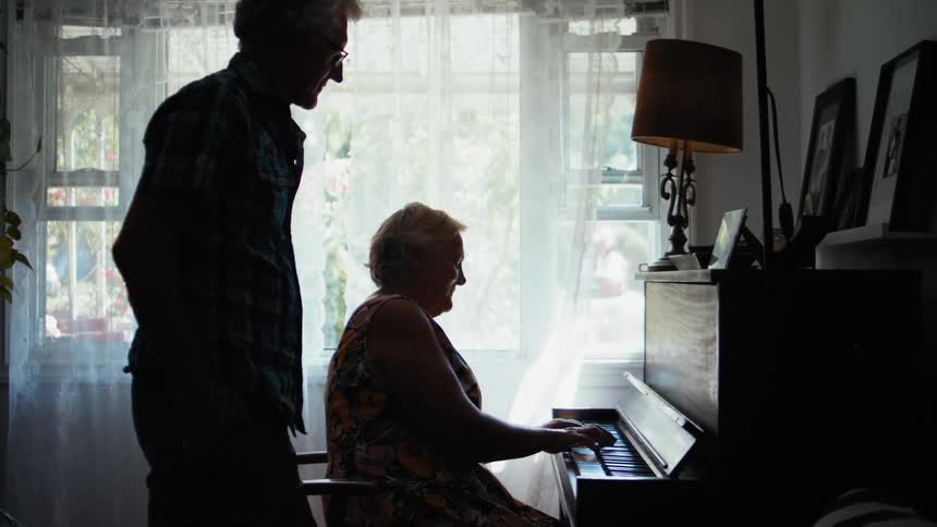 Elderly man standing next to woman playing the piano is silhouetted against windows