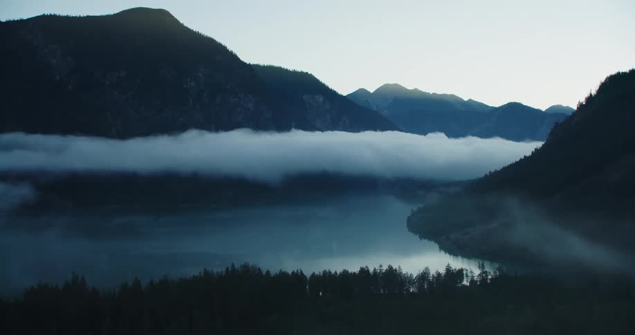 Fog covers conifer forest bordering lake between mountains separated by low lying cloud