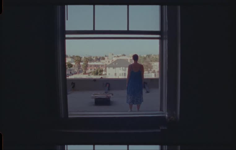 Through a window a woman can be seen standing motionlessly on the rooftop of a building in a city neighborhood