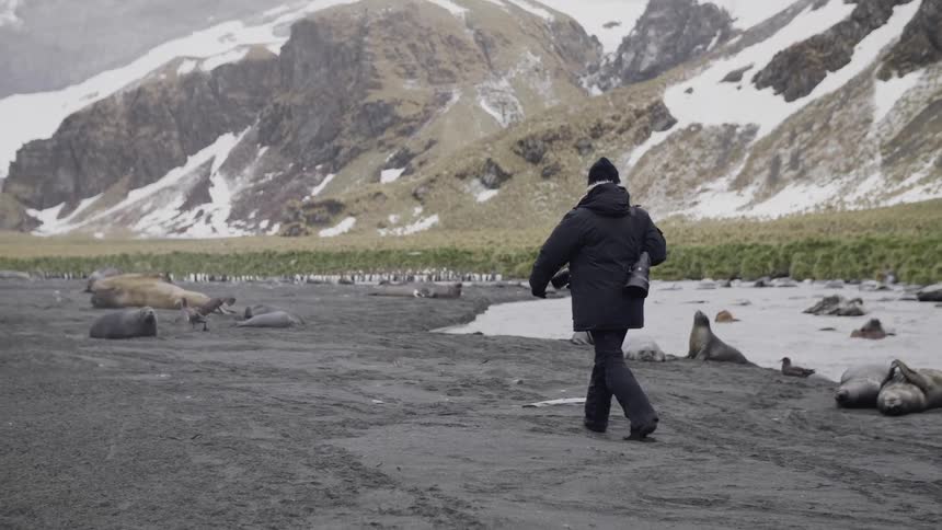 A person walks across a dirt terrain beside a small creek and large snowy mountains