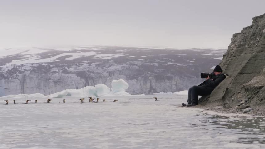 A man in warm clothing with a camera is laying against a moutain side shooting pictures of penguins swimming in the ice filled ocean