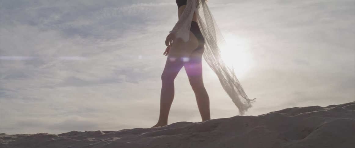 A white woman in a black bikini and sheer wrap walks across the tan sand