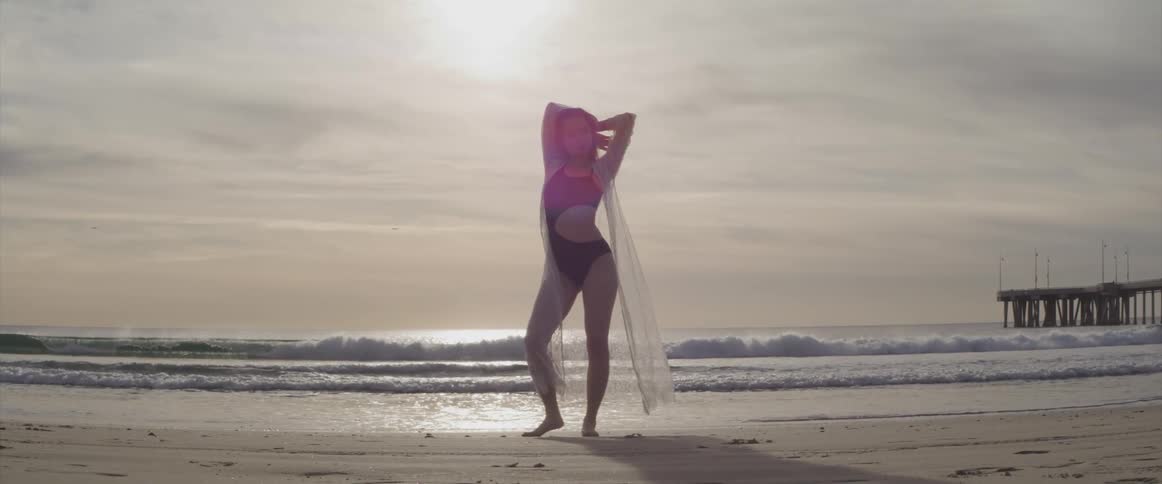Woman wearing swimsuit and wrap poses on beach back lit by sun in cloudy sky