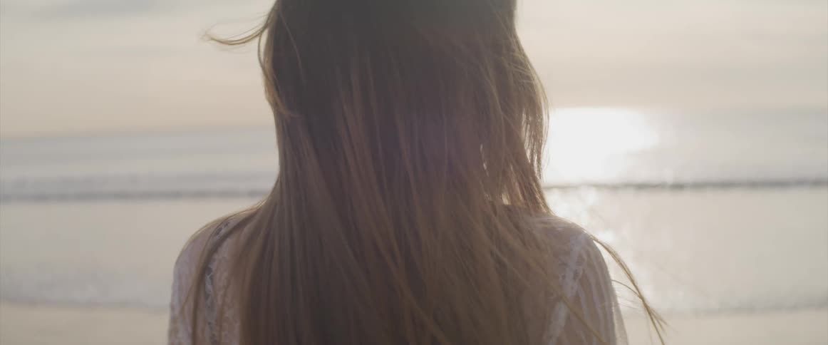 A young asian woman raising looks into the camera before slowly turning around to gaze at the ocean