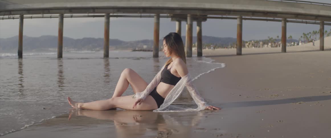 A young asian woman in a swimsuit sits in the sand on the beach as the tide washes in around her