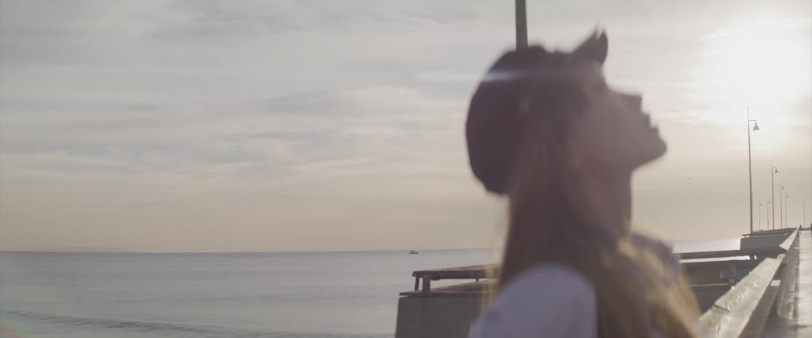 A woman with a hat on a pier looking up at the sky with the ocean and the sun in the background