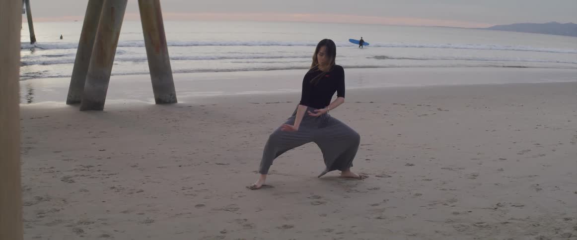 Woman performs tai chi on the beach while surfer wades through surf as sky on horizon is tinted red