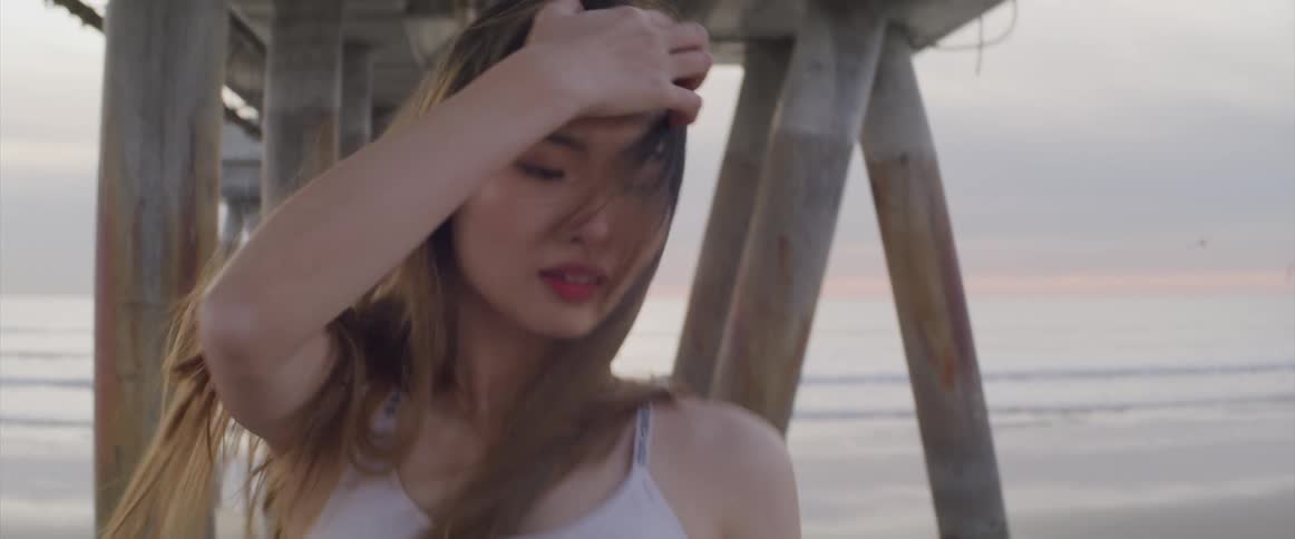 Young woman dances looking at the camera under a pier on the beach at sunset