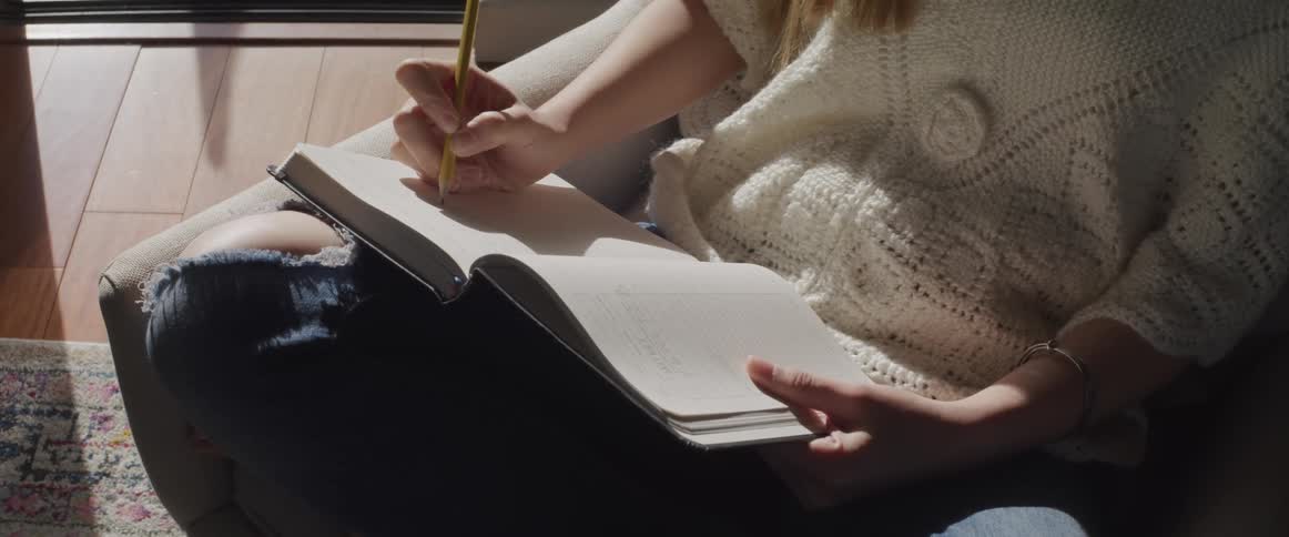 Woman with ripped jeans and a white sweater sitting in an armchair writes things down in a notebook