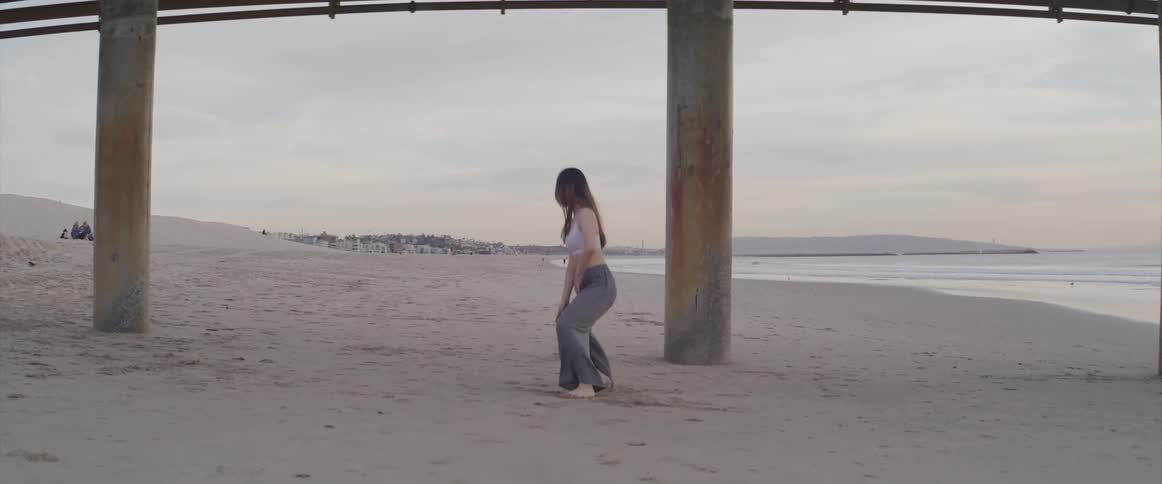 A woman does tai chi under the pier on a beach with a dense neighborhood in the distance
