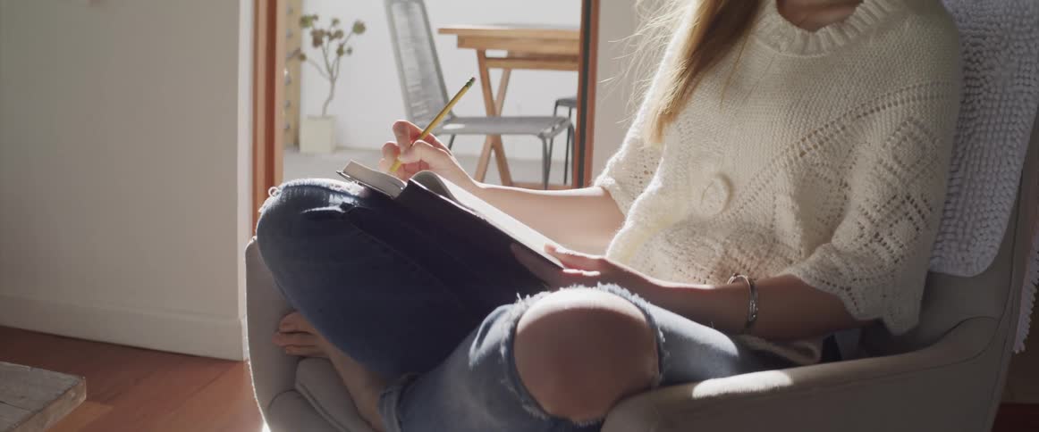 Woman with ripped jeans and a white sweater sits in an armchair and writes things down in a notebook