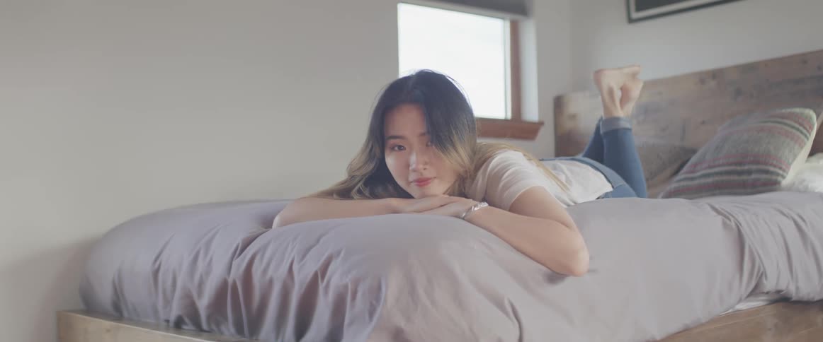 Asian woman lies face down on a bed and looks at the camera in the bedroom