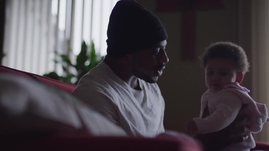 Black man with a beanie and a white sweatshirt sits on a red sofa and holds his toddler girl in the living room at home