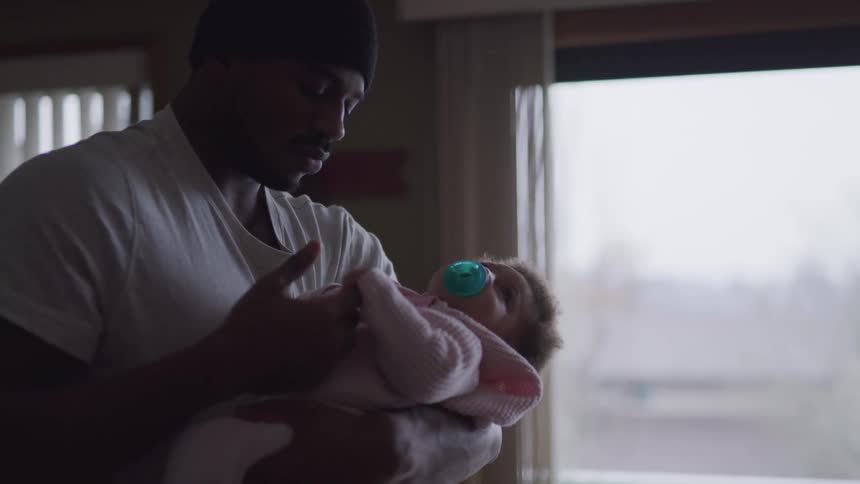 A man gently rocks a baby as it gets one of his fingers while standing near a window in a house during the day