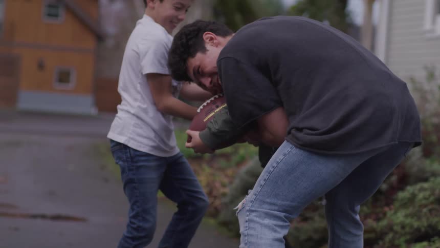 An adult male playfully wraps his arms around a younger boy carrying a football