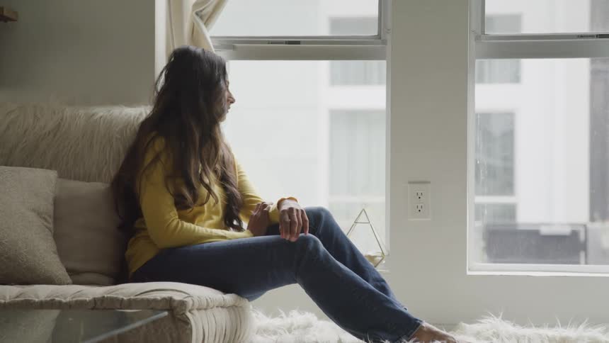Woman sits on couch looking out window at opposite building