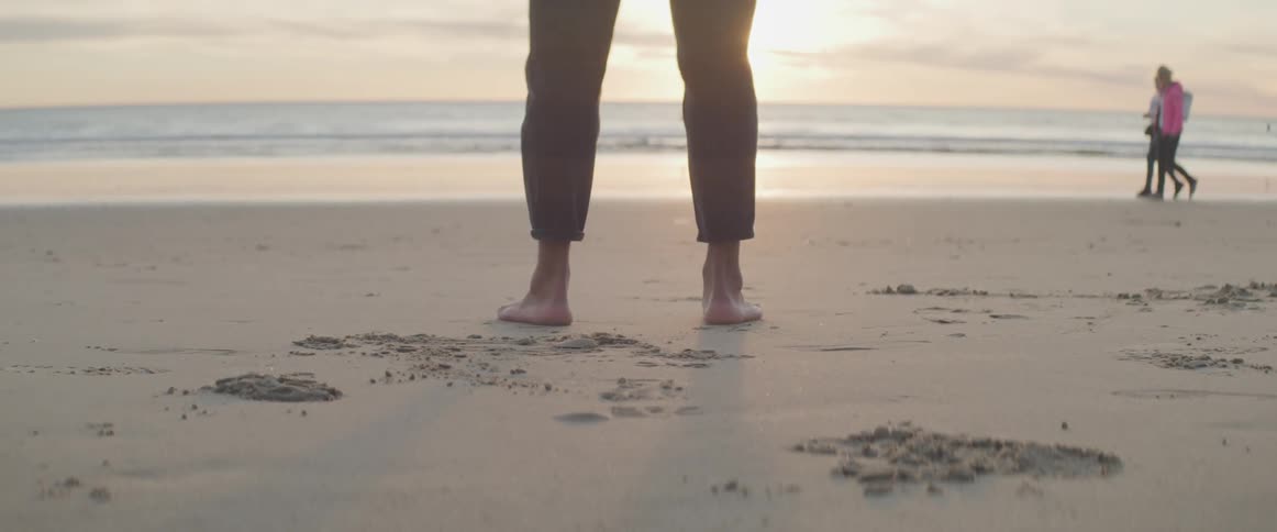 Person stands facing sea as people walk past near the surf