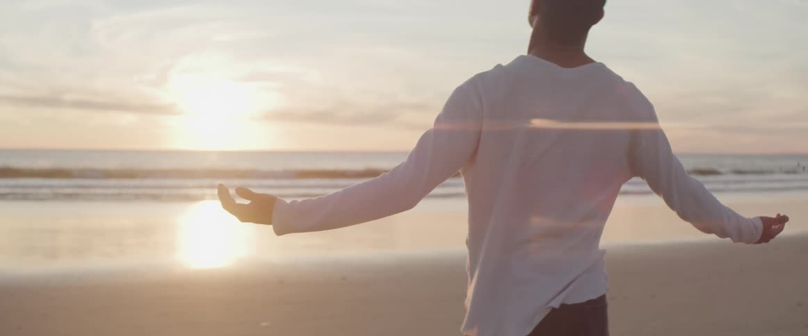 Young man stands on the sand beach with arms wide open at sunset