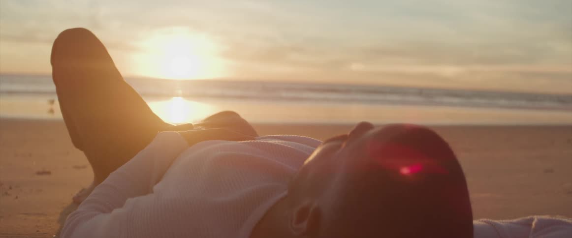 Black man with a white sweatshirt and black pants lies on the beach next to the sea at sunset