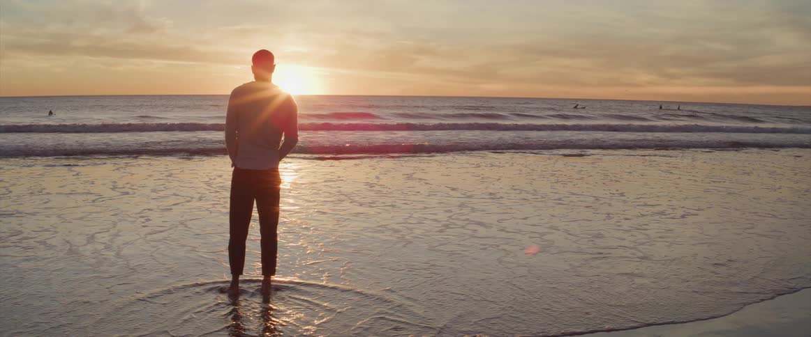 A man stands on a beach shore looking out at the ocean and sunset
