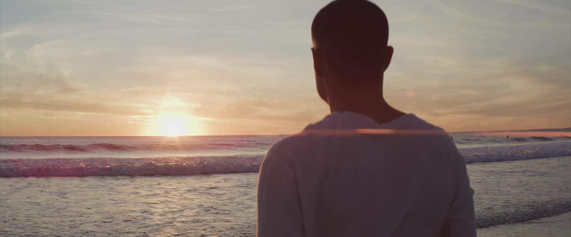 A man standing on a beach looks out over the ocean towards the horizon as the sun sets