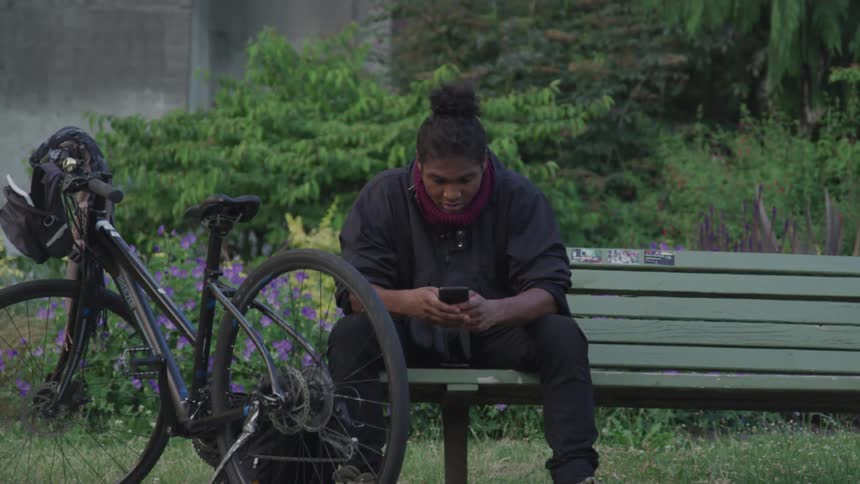 A teenage boy sits on a bench next to his bike and texts on his cell phone