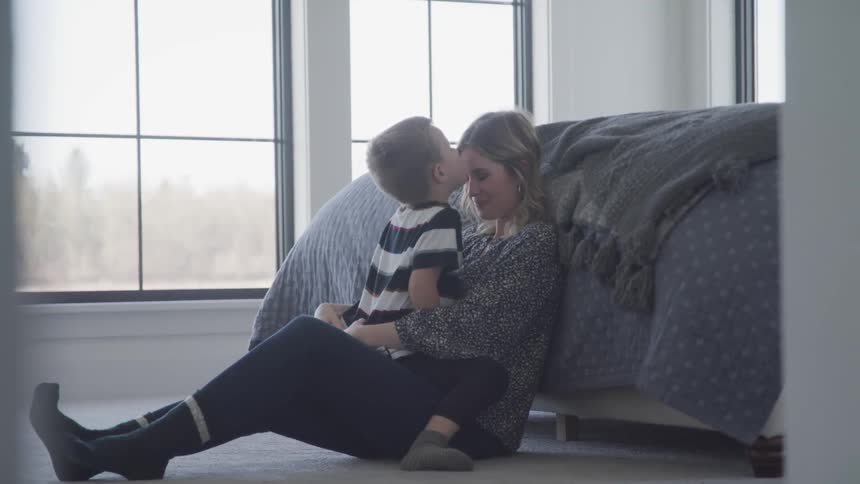 A young boy in a striped shirt sits in his mothers lap and kisses her forehead as they rest on the ground against a bed in an upstairs bedroom