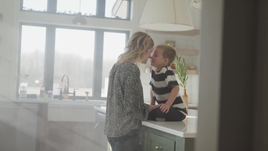 A woman giving eskimo kisses to a boy sitting on a counter in a kitchen