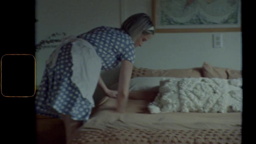 Blonde housewife with a blue dress makes the bed in bedroom